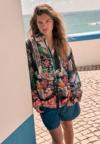 Kleurrijk bloemetjesoverhemd met lange mouwen, gecombineerd met denim shorts; model leunt tegen een muur en glimlacht.