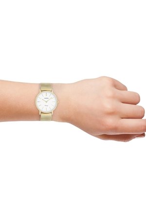 Montre bracelet en maille dorée avec un cadran blanc, affichant de fins marqueurs horaires et un design minimaliste. Portée sur un poignet à la peau claire.