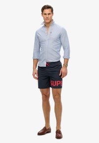 Camisa de botones a rayas azul claro, pantalones cortos de baño azul marino con gran impresión roja de "SUPER", mocasines marrones y un reloj de pulsera.