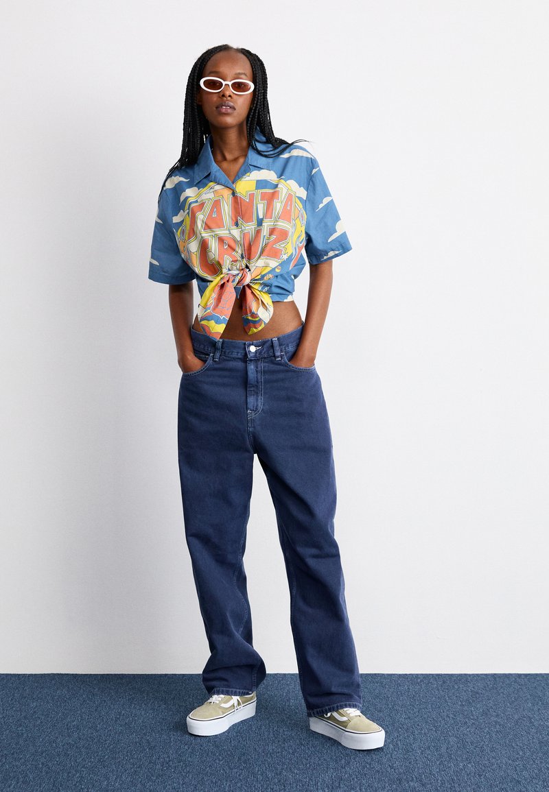 Camisa gráfica azul con un nudo en el frente, combinada con pantalones vaqueros sueltos de color azul oscuro y zapatillas beige con acentos blancos. Se llevan gafas de sol blancas.