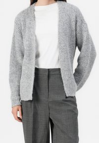 Femme portant un cardigan gris en tricot par-dessus une chemise blanche et un pantalon gris à carreaux taille haute, sur un fond uni.