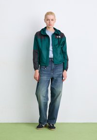Teal windbreaker jack met grijze mouwen, verstelbare capuchon, ritszakken en elastische manchetten. Gecombineerd met wijde blauwe jeans en zwarte sneakers.