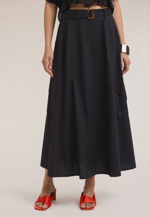 Jupe longue noire avec une taille ceinturée, dotée de poches latérales et d'une texture lisse. Associée à des talons ouverts rouges.