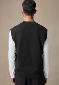 Schwarzer gestrickter ärmelloser Pullover mit geripptem Halsausschnitt und Saum, getragen über einem langärmeligen weißen Hemd. Weiche Textur, schlichtes Design.