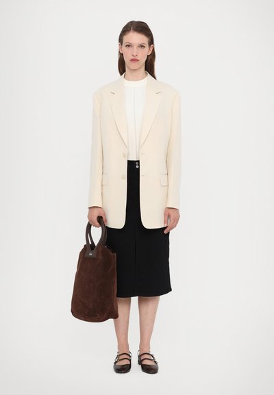 Blazer crema sopra top bianco, gonna nera, che trasporta una borsa a tracolla in suede marrone con manico; scarpe scure; design minimalista, texture lisce.