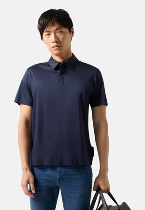 Man draagt een marineblauw poloshirt met korte mouwen en spijkerbroek, houdt een zwarte leren tas vast tegen een effen lichte achtergrond.