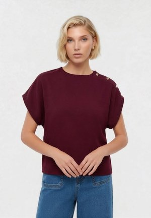 Femme blonde portant un haut bordeaux à manches courtes avec des boutons sur l'épaule et un jean bleu, debout les mains jointes.