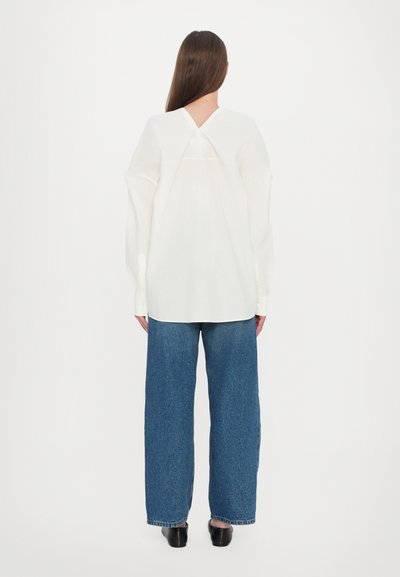 Witte blouse met lange mouwen en een V-vormige achterkant, gecombineerd met brede spijkerbroek in blauw. De stof lijkt lichtgewicht en iets doorschijnend te zijn.