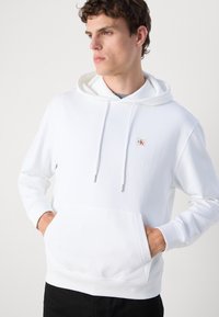 Jaunas vyras dėvi baltą gobtuvą su priekiniu kišeniu ir mažu „ck“ logotipu ant krūtinės, rankos kišenėse, žiūri į šoną.