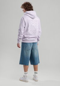 Vaaleanvioletissa hupparissa on valkoinen graafinen kuvio selässä, joka on yhdistetty sinisiin farkkushortsihin ja valkoisiin tennareihin, joissa on vihreät raidalliset sukat.