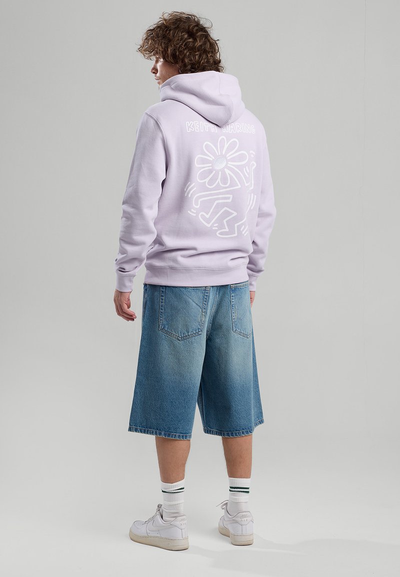 Vaaleanvioletissa hupparissa on valkoinen graafinen kuvio selässä, joka on yhdistetty sinisiin farkkushortsihin ja valkoisiin tennareihin, joissa on vihreät raidalliset sukat.