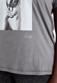 Harmaa puuvillapaita, jossa on graafinen painatus kädestä, joka pitää helmiäkoruja, ja "vibes" brodeerattuna hopealla. Pyöreä kaula-aukko ja lyhyet hihat.