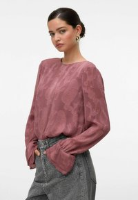 Blusa a maniche lunghe in mauve con motivo floreale testurizzato; presenta un colletto rotondo e polsini svasati, abbinata a jeans grigi a vita alta con motivo.