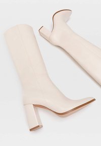 Bottes en cuir blanc montantes, au design élégant, avec des talons carrés et une texture lisse. Présentent un éclat subtil et une semelle marron pour le contraste.