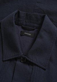 Tumši zila kokvilnas jaka ar apkakli, vienu melnu pogu aizdarē un zīmola etiķeti. Raksturo teksturēta auduma un tīrs dizains.