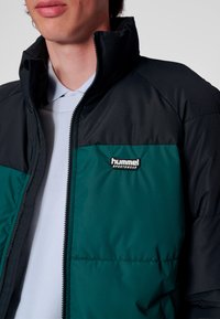 Kurtka ocieplana z czarną górną i turkusową dolną częścią. Posiada wysoki kołnierz, zamek błyskawiczny oraz logo 'hummel Sportswear' na piersi.