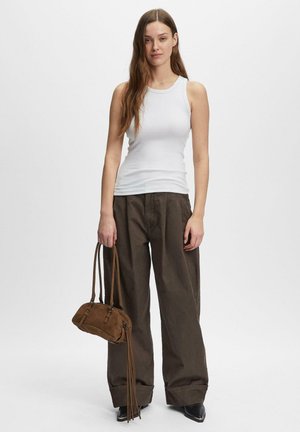 Młoda kobieta z długimi włosami, ubrana w biały top bez rękawów, szerokie brązowe spodnie, czarne buty, trzyma brązową torebkę z frędzlami.