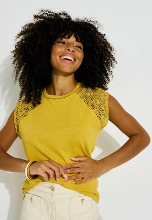 Mujer con pelo rizado sonríe, lleva una blusa amarilla con mangas de encaje y pantalones de color claro, posando frente a un fondo blanco liso.