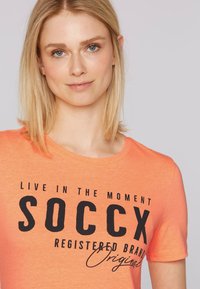 T-shirt arancione a maniche corte con scollatura rotonda. Presenta un testo nero in grassetto che recita "VIVI NEL MOMENTO SOCCX MARCHIO REGISTRATO Originale."