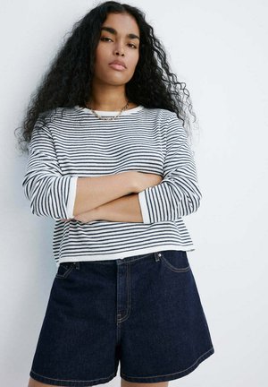 Femme aux longs cheveux bouclés porte un t-shirt à manches longues rayé blanc et noir et un short en jean foncé, bras croisés sur fond blanc.