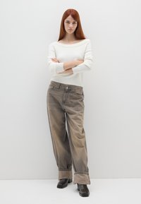 Weißer Off-Shoulder-Pullover, lockere beige Jeans mit umgekrempelten Säumen und schwarze Absatzstiefel. Das Model steht mit verschränkten Armen.