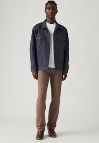 Mørkeblå denimjakke med brystlommer og kobberfarvet syninger, båret over en hvid t-shirt, parret med lysebrune bukser og brune støvler.