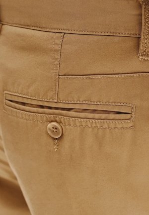Beżowe spodnie z tylną kieszenią z poziomymi szczelinami i zapięciem na guzik. Tkanina ma gładką fakturę i subtelny połysk.