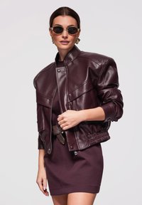 Femme portant une veste en cuir bordeaux foncé sur une robe assortie, accessoirisée d'une ceinture marron, de boucles d'oreilles dorées et de lunettes de soleil, sur un fond clair.