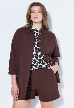 Kobieta ubrana w brązową koszulę zapinaną na guziki z podwiniętymi rękawami, pasujące szorty oraz biały top w czarne i brązowe abstrakcyjne plamy.