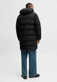 Schwarze gepolsterte Jacke mit Kapuze, ausgestattet mit einem gesteppten Design und langer Silhouette. Kombiniert mit weiten blauen Jeans und schwarzem Schuhwerk.