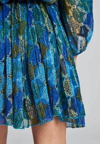 Falda plisada en azul y verde con patrones florales. Tejido traslúcido con acentos dorados que muestra textura y movimiento. Los bordes son ondulados.
