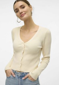 Krämfärgad ribbad långärmad cardigan med V-halsringning och framknäppning, kombinerad med ljusblå denimjeans.