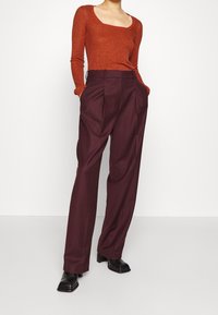 Femme portant un top à manches longues en maille couleur rouille, un pantalon prune taille haute ample et des bottes à talons noires à bout carré, les mains dans les poches.