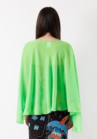 Blusa trasparente verde con maniche lunghe e larghe e scollatura rotonda, leggera e sovrapposta, indossata sopra pantaloni neri a fantasia.