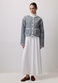 Prešívaná sivá bunda s bielymi lemami a gombíkmi, vybavená veľkými vreckami. Nosená cez dlhé biele šaty a hnedé mokasíny.