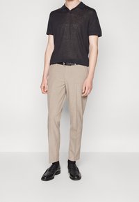 Zwarte polo met korte mouwen, lichtbeige op maat gemaakte broeken, zwarte leren instapschoenen en zwarte sokken, gepresenteerd in een staande pose.