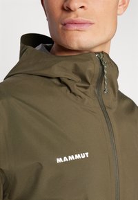 Maslinasto zelena vodootporna jakna s patentnim zatvaračem, visokim ovratnikom i bijelim logotipom "MAMMUT" na prsima. Glatka tekstura, lagana konstrukcija.