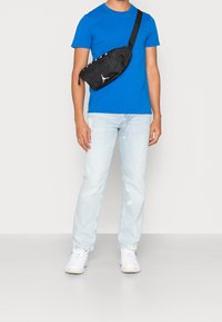 Černá ledvinka s hladkou texturou, vybavená bílým logem a nastavitelným popruhem, nošená přes jasně modré tričko a světlé džíny.