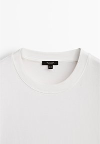 T-shirt bianco in cotone con collo rotondo ribattuto, cuciture piatte e una piccola etichetta nera all'interno del colletto che indica la taglia M.