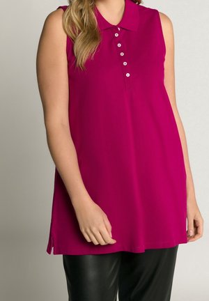 Polo sans manches magenta avec six boutons blancs porté par une personne aux longs cheveux blonds, associé à un pantalon noir.