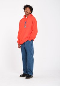 Een oranje hoodie met een voorzak en grafisch ontwerp, gecombineerd met blauwe spijkerbroeken en zwarte schoenen. Modell staat tegen een witte achtergrond.