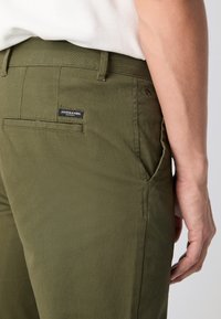 Olivegrønne bukser med lige snit, sidelommer og en baglomme med en sort label. Glat tekstur og robust stof.