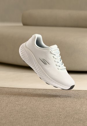 Bela športna superga s teksturiranim podplatom in mrežastim zgornjim delom, z srebrnim logotipom "S", ki lebdi nad bež preprogo in ozadjem kavča.