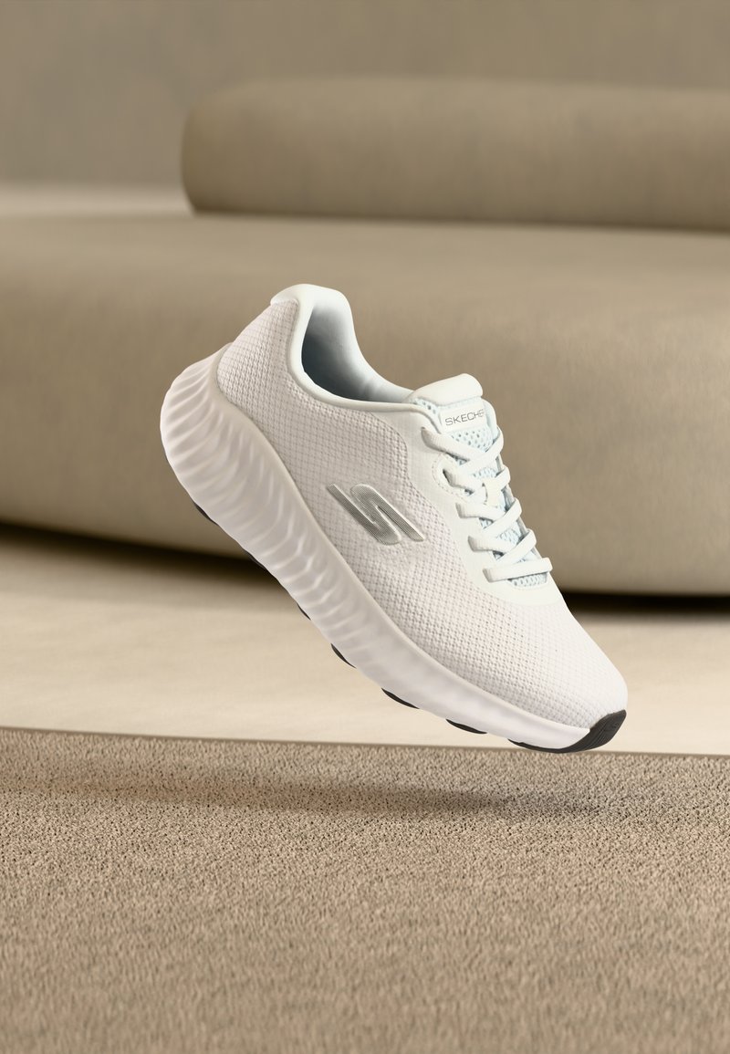 Λευκό αθλητικό παπούτσι με υφασμένο πέλμα και επάνω μέρος από διχτυωτό υλικό, με ασημί λογότυπο "S", αιωρούμενο πάνω από μια μπεζ μοκέτα και φόντο με καναπέ.