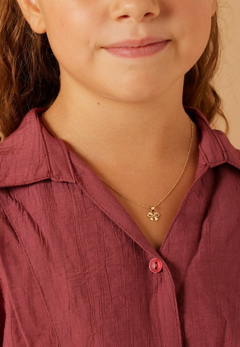 Goldener Blumenanhänger mit einer zarten Kette, getragen über einem kastanienbraunen strukturierten Shirt mit einer rosa Knopf. Nahaufnahme des Ausschnitts.