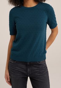 Laureat sweter z krótkim rękawem w kolorze zielono-niebieskim, z fakturowanym wzorem kropek oraz prążkowanym dekoltem i dolnym wykończeniem. Połączony z ciemnoszarymi dżinsami.