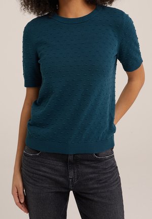 Suéter de punto de manga corta en color verde azulado, con un patrón de puntos texturizados y cuello y dobladillo acanalados. Combinado con jeans de lona oscura.