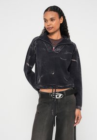 Samazināts melns fliša pulovers ar teksturētu apdari, puszipa apkakli un regulējamu apakšu ar aukliņu. Kombinēts ar tumšām platajiem džinsiem un melnu jostu.