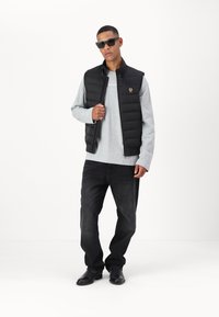 Black padded vest with horizontal stitching, grey long-sleeve shirt, black jeans, and black shoes; model wearing sunglasses.