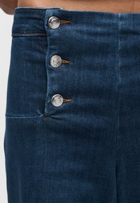 Κοντινό πλάνο σκουρόχρωμης μπλε τζιν φούστας με τρία ασημένια κουμπιά με την ετικέτα "ONLY" και πορτοκαλί ραφές στην αριστερή πλευρά.